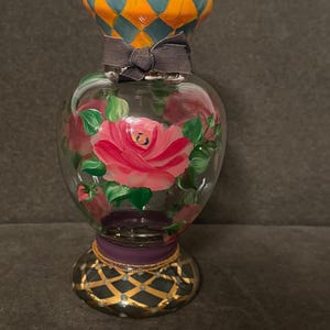 May include: A clear glass vase featuring hand-painted pink roses with green leaves. The neck has an orange and teal diamond pattern, a dark gray ribbon tied in a bow, and the base is purple with a black and gold lattice design.