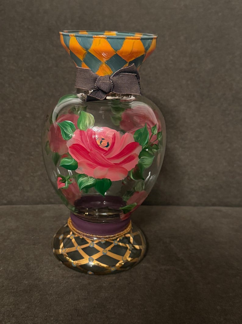 May include: A clear glass vase featuring hand-painted pink roses with green leaves. The neck has an orange and teal diamond pattern, a dark gray ribbon tied in a bow, and the base is purple with a black and gold lattice design.