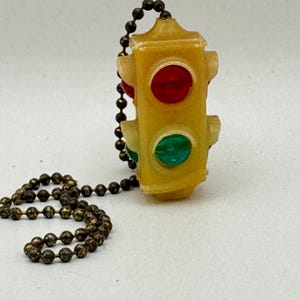 May include: A yellow traffic light charm with red and green lights, attached to a bronze-colored ball chain. The charm is a miniature replica of a traffic signal, with a rectangular shape and rounded edges.