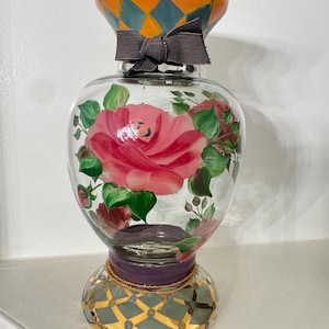 May include: A clear glass vase featuring hand-painted pink roses with green leaves on the body. The neck has an orange and teal diamond pattern with a dark gray fabric bow. The base is decorated with a gold and teal grid pattern and a purple band.
