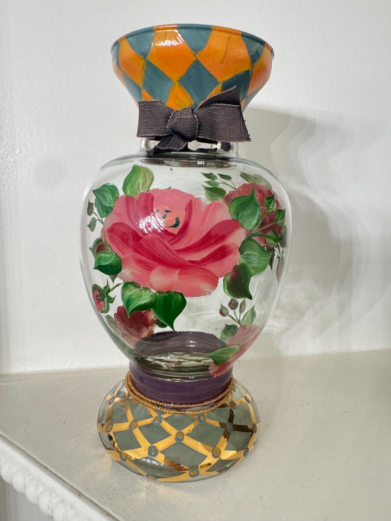 May include: A clear glass vase featuring hand-painted pink roses with green leaves on the body. The neck has an orange and teal diamond pattern with a dark gray fabric bow. The base is decorated with a gold and teal grid pattern and a purple band.