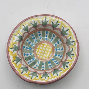 May include: A round, hand-painted ceramic bowl with a colorful design. The rim is a muted pink, with a yellow and green floral pattern. The center features a yellow grid pattern surrounded by blue dots and green shapes.