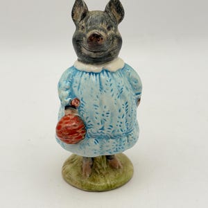 May include: A ceramic figurine of a pig standing on a circular green base. The pig has a dark grey head and is wearing a light blue dress with a white collar. The pig is holding a small, round, textured red object in its right hand.