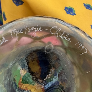 May include: A clear glass object with a rounded base featuring an etched signature that reads MacKenzie-Childs 1997. The glass contains visible air bubbles and a multi-colored interior pattern with shades of green, blue, red, and yellow.
