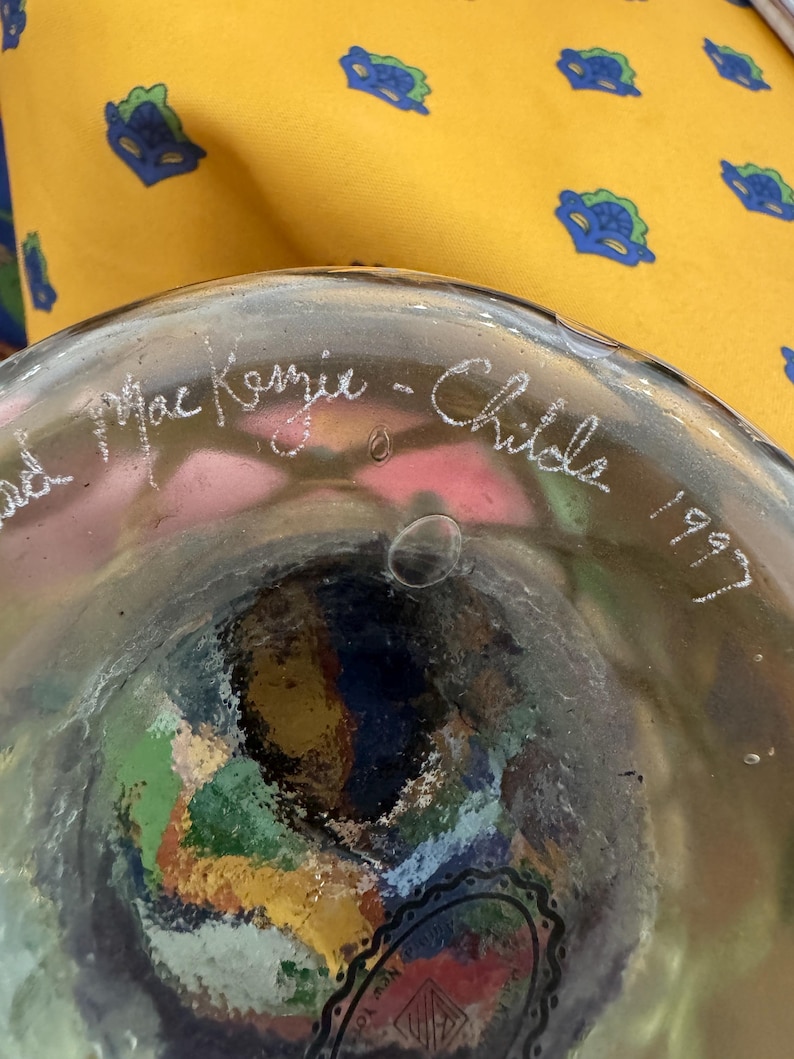 May include: A clear glass object with a rounded base featuring an etched signature that reads MacKenzie-Childs 1997. The glass contains visible air bubbles and a multi-colored interior pattern with shades of green, blue, red, and yellow.