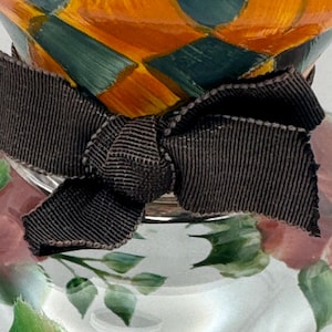 May include: A glass vessel featuring a hand-painted diamond pattern in orange and teal at the top, accented by a dark brown grosgrain ribbon tied in a bow around the neck, with a floral design painted in pink and green on the clear glass base.
