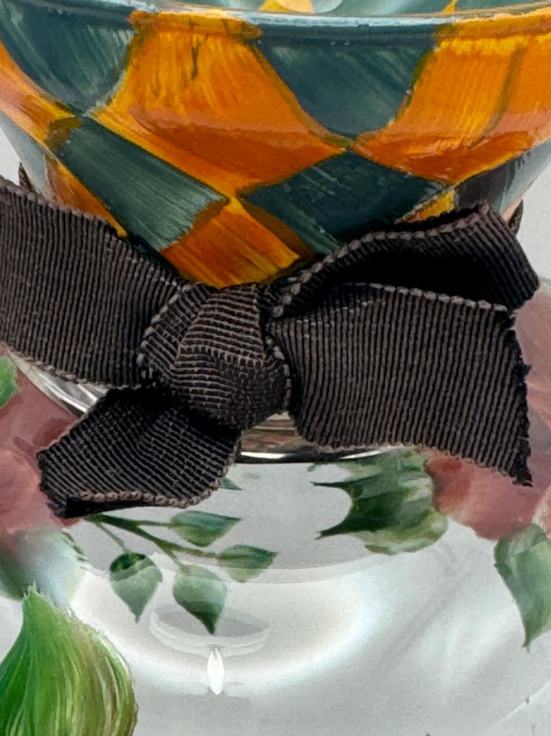 May include: A glass vessel featuring a hand-painted diamond pattern in orange and teal at the top, accented by a dark brown grosgrain ribbon tied in a bow around the neck, with a floral design painted in pink and green on the clear glass base.
