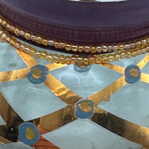 May include: A glass vessel featuring a light blue and gold diamond pattern with small blue and gold circular accents. Two strands of iridescent amber-colored seed beads are wrapped around the neck of the glass container.