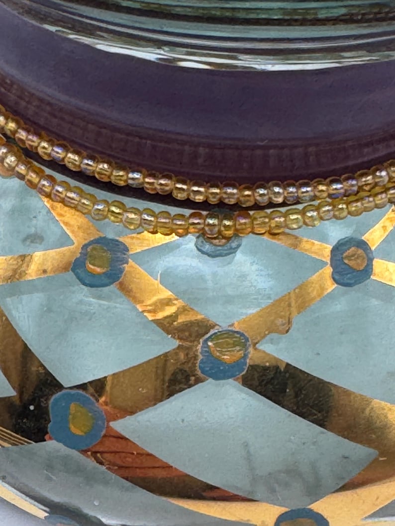 May include: A glass vessel featuring a light blue and gold diamond pattern with small blue and gold circular accents. Two strands of iridescent amber-colored seed beads are wrapped around the neck of the glass container.