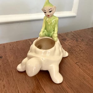 May include: A ceramic planter shaped like a white turtle with a glossy finish. A small, light green elf figure with pointed ears, dark eyebrows, and red lips is attached to the top of the turtle shell, which features a circular opening for plants.