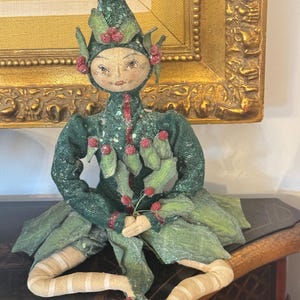 May include: A seated, handmade elf doll with a whimsical design. The doll features a green outfit with leaf and berry accents, a pointed hat, and striped stockings. The doll's face is painted with delicate features, and it is posed on a dark wooden surface.