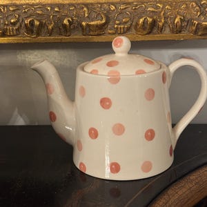 May include: A white ceramic teapot with a lid, adorned with pink polka dots. The teapot features a spout, handle, and a small knob on the lid. The teapot is sitting on a dark surface.