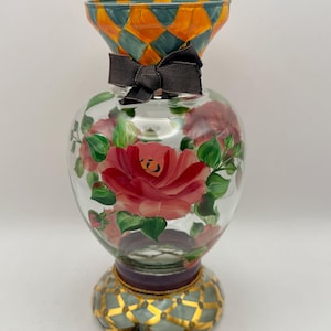 May include: A clear glass vase featuring hand-painted pink roses with green leaves on the body. The flared rim and the circular base are decorated with an orange and teal diamond pattern. A dark brown fabric ribbon is tied around the neck.