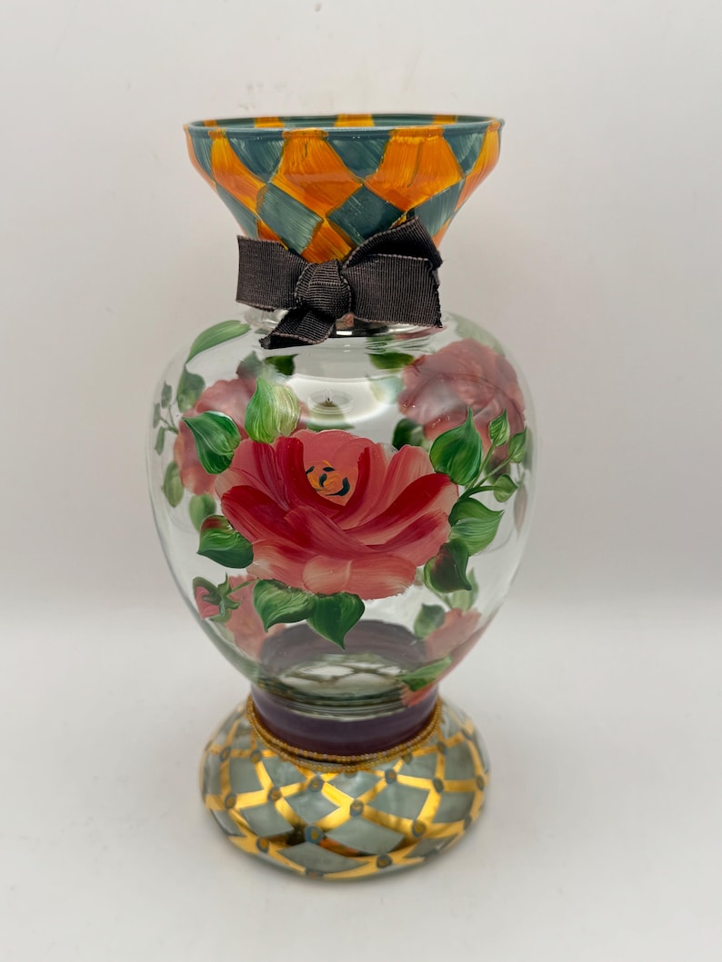 May include: A clear glass vase featuring hand-painted pink roses with green leaves on the body. The flared rim and the circular base are decorated with an orange and teal diamond pattern. A dark brown fabric ribbon is tied around the neck.