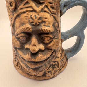 May include: Handmade ceramic mug with a smiling face design. The mug features a brown exterior with carved details and a blue handle and rim. The face has detailed eyes, nose, and mouth, with star and geometric patterns.
