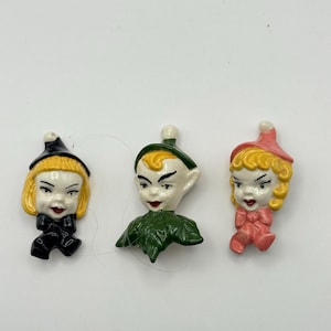 May include: Three vintage ceramic figurines. One is a witch with a black hat and outfit, another is a green-clad elf, and the third is a girl in a pink outfit and hat. Each figurine has a painted face with red lips and detailed features.