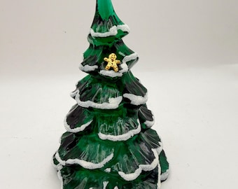 May include: A translucent green glass Christmas tree figurine featuring textured white snow detailing on the edges of its branches and a small, gold-colored gingerbread man ornament attached to the front of the tree near the top.