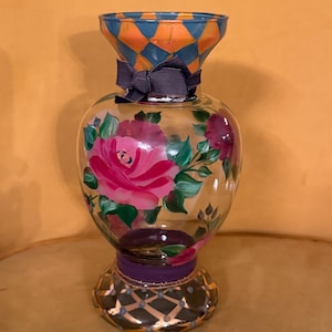 May include: A clear glass vase featuring a hand-painted pink rose design with green leaves. The neck has an orange and blue diamond pattern, while the base displays a dark purple band and a gold-accented grid. A dark gray ribbon is tied around the neck.