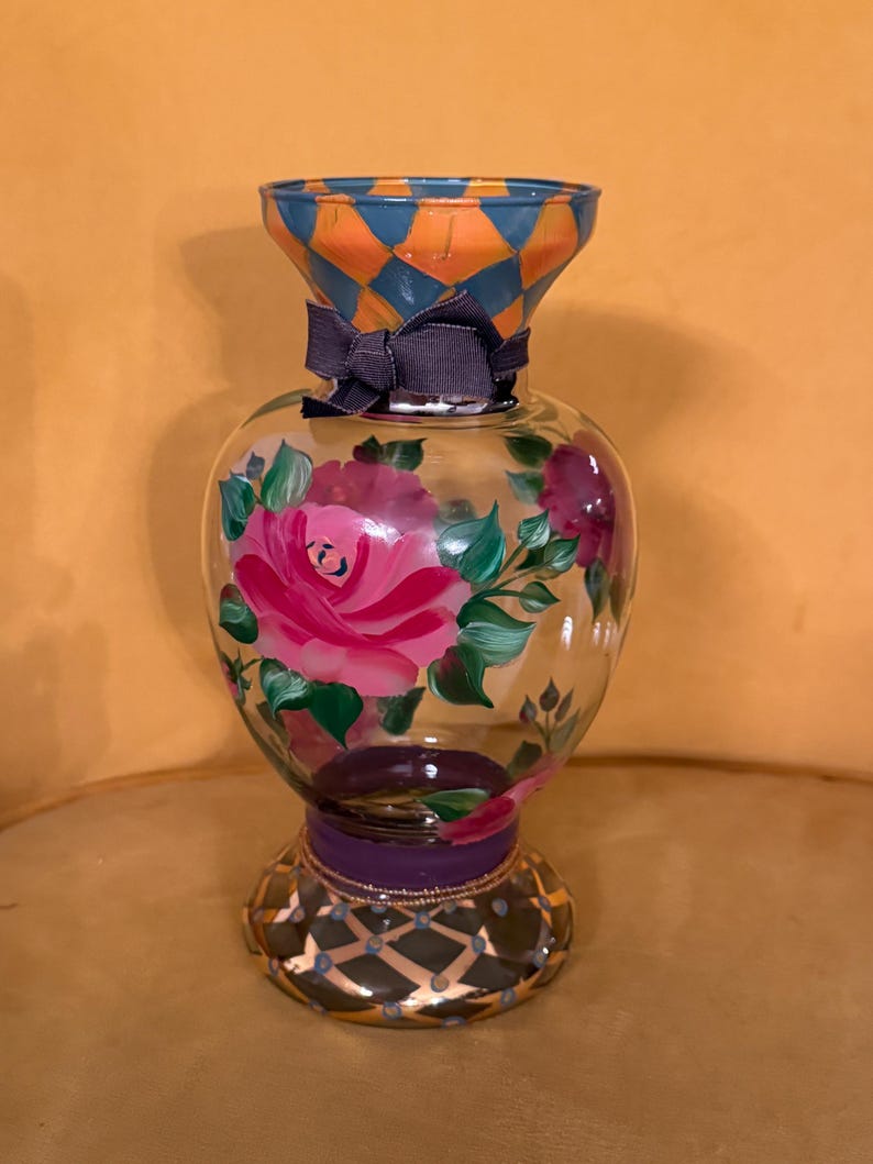 May include: A clear glass vase featuring a hand-painted pink rose design with green leaves. The neck has an orange and blue diamond pattern, while the base displays a dark purple band and a gold-accented grid. A dark gray ribbon is tied around the neck.