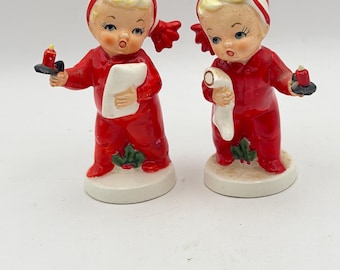 Może przedstawiać: Dwie ceramiczne figurki dzieci w czerwonych piżamach i czerwono-białych czapkach nocnych. Każda postać trzyma mały czarny świecznik z czerwoną świecą i biały zwój, stojąc na okrągłych białych podstawach z zielonymi akcentami ostrokrzewu.