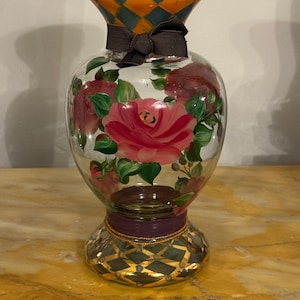 May include: A clear glass vase featuring hand-painted pink roses with green leaves on the body. The neck and base are decorated with an orange and teal diamond pattern. A dark brown ribbon is tied around the neck, and a purple band sits above the base.