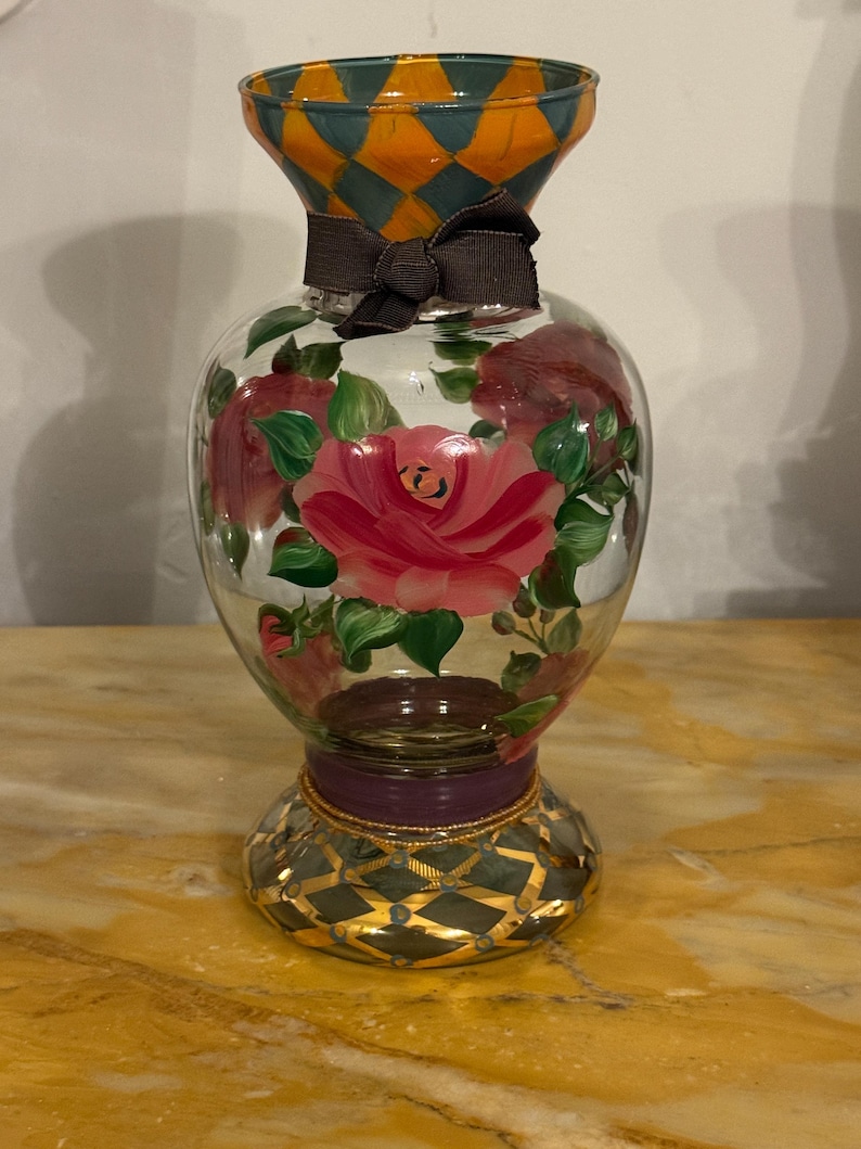 May include: A clear glass vase featuring hand-painted pink roses with green leaves on the body. The neck and base are decorated with an orange and teal diamond pattern. A dark brown ribbon is tied around the neck, and a purple band sits above the base.