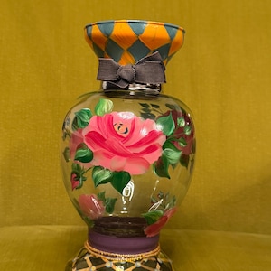May include: A clear glass vase featuring hand-painted pink roses with green leaves on the body. The neck has an orange and blue diamond pattern, while the base displays a black and gold grid design. A dark gray ribbon is tied around the neck.
