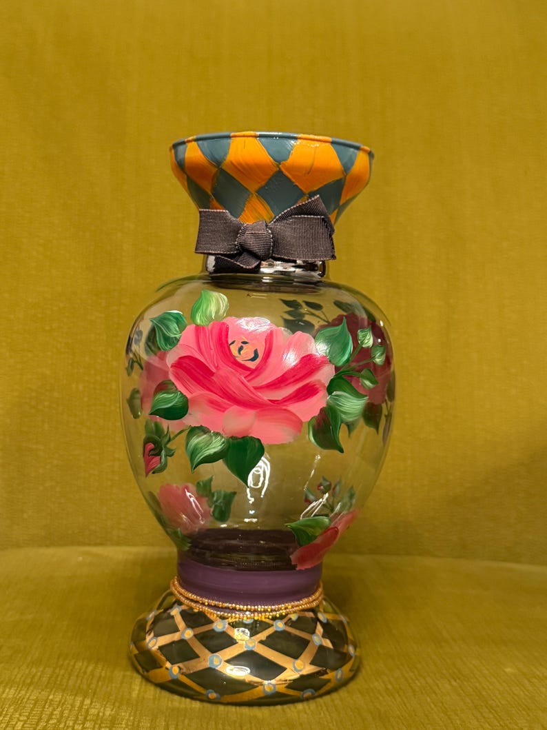 May include: A clear glass vase featuring hand-painted pink roses with green leaves on the body. The neck has an orange and blue diamond pattern, while the base displays a black and gold grid design. A dark gray ribbon is tied around the neck.