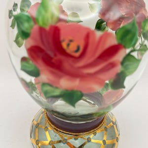 May include: A clear glass perfume bottle featuring hand-painted pink roses and green leaves. The base is decorated with a gold lattice pattern over a light blue background, accented with small blue dots and a gold beaded trim at the neck.