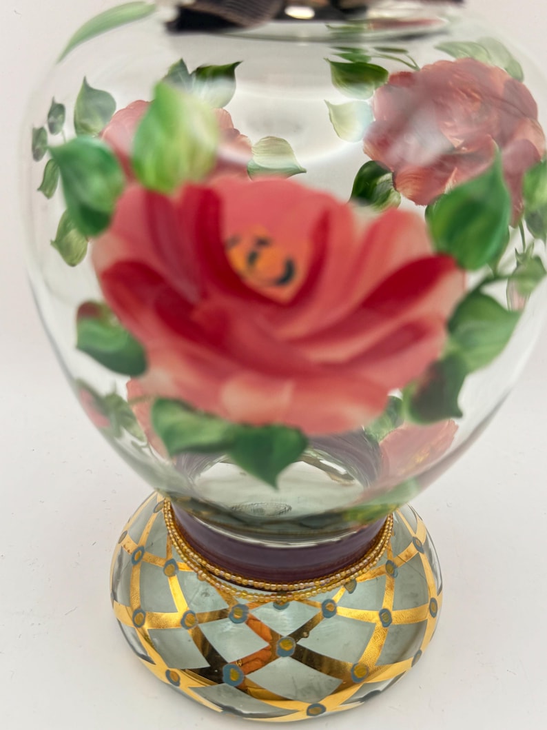 May include: A clear glass perfume bottle featuring hand-painted pink roses and green leaves. The base is decorated with a gold lattice pattern over a light blue background, accented with small blue dots and a gold beaded trim at the neck.
