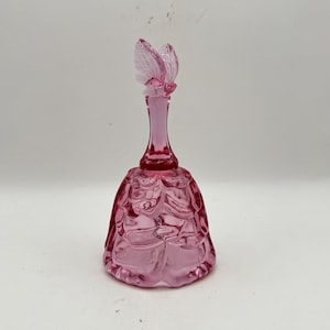 May include: A pink glass bell-shaped perfume bottle with a butterfly stopper. The bottle has a detailed, textured design and a long, slender neck. The butterfly stopper adds a decorative touch to the vintage-style bottle.