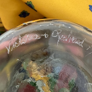 May include: A clear glass object with an etched inscription reading Victoria to Richard. The glass features a textured, multi-colored interior with visible red, yellow, and green tones. A yellow fabric with a blue and green pattern is visible.