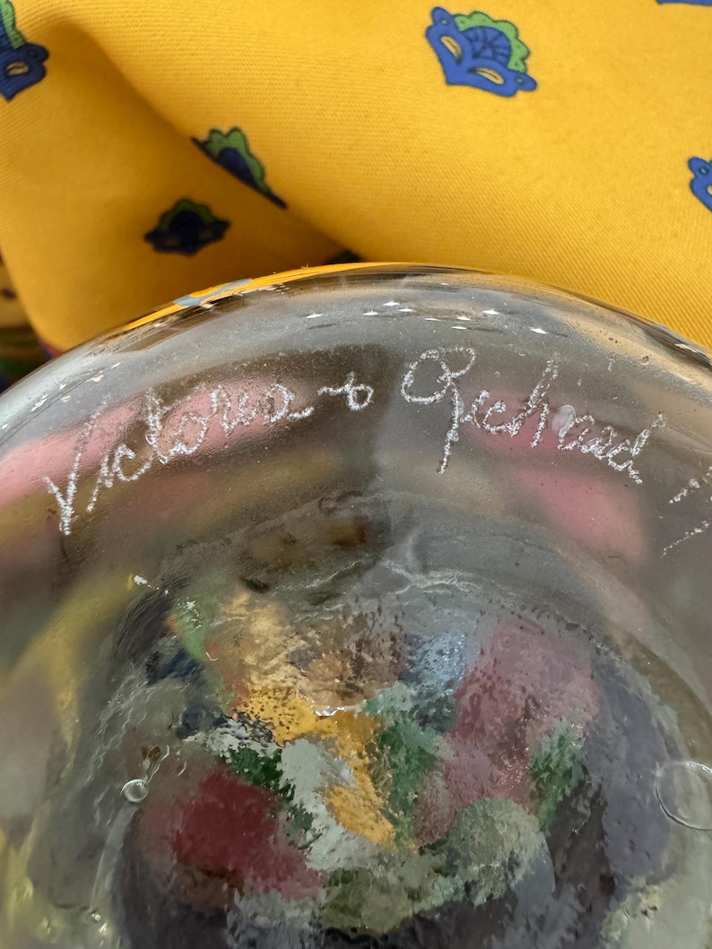 May include: A clear glass object with an etched inscription reading Victoria to Richard. The glass features a textured, multi-colored interior with visible red, yellow, and green tones. A yellow fabric with a blue and green pattern is visible.