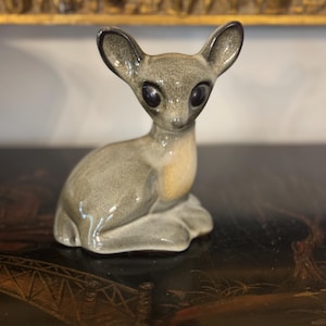May include: A ceramic figurine of a deer in a seated position. The deer has large, dark eyes, long ears, and a gray body with a lighter belly. The figurine is on a dark surface, and the background includes a gold-colored decorative frame.