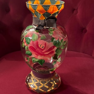 May include: A clear glass vase featuring a hand-painted pink rose design with green leaves. The neck has an orange and dark gray diamond pattern, while the base displays a dark gray lattice design with gold accents. A dark brown ribbon is tied around the neck.