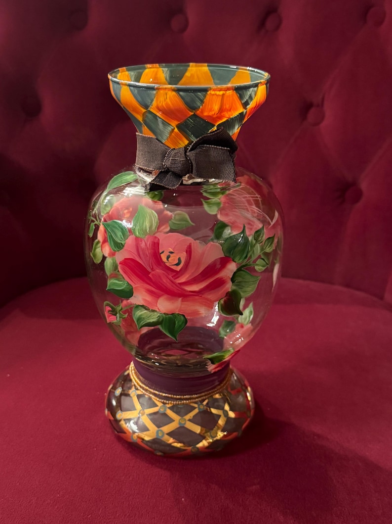 May include: A clear glass vase featuring a hand-painted pink rose design with green leaves. The neck has an orange and dark gray diamond pattern, while the base displays a dark gray lattice design with gold accents. A dark brown ribbon is tied around the neck.