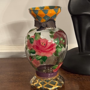 May include: A clear glass vase featuring hand-painted pink roses with green leaves on the body. The neck and base are decorated with an orange and teal diamond pattern. A dark gray ribbon is tied around the neck, and a purple band accents the base.