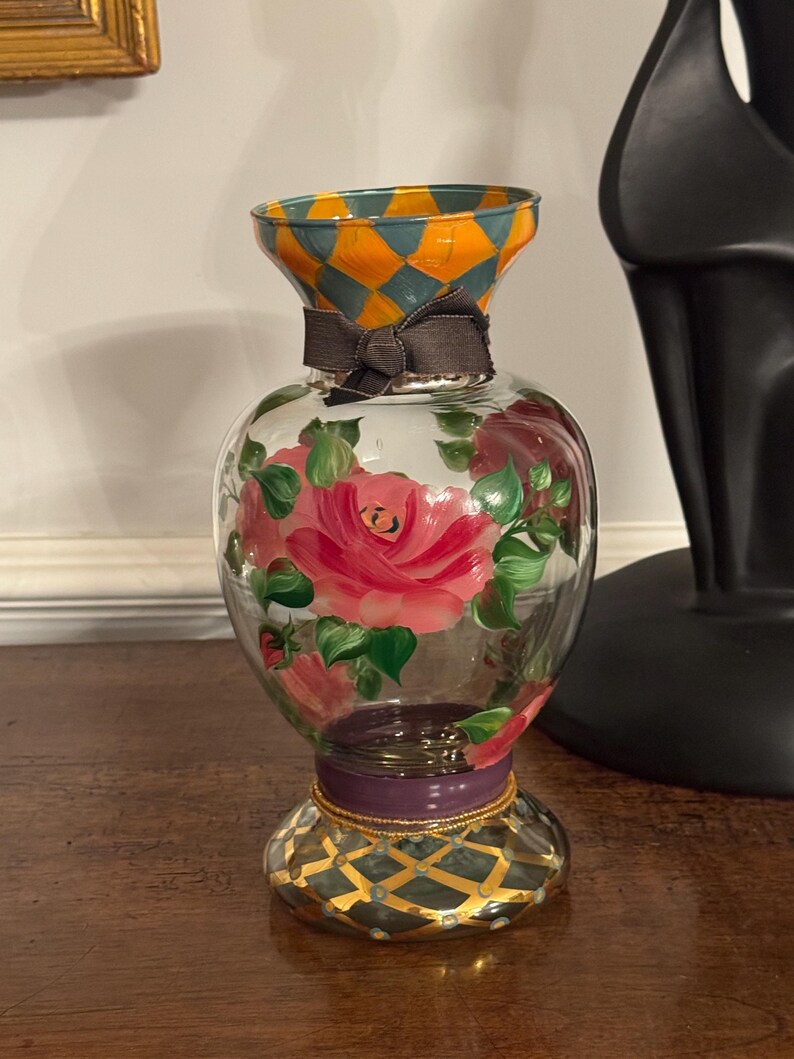 May include: A clear glass vase featuring hand-painted pink roses with green leaves on the body. The neck and base are decorated with an orange and teal diamond pattern. A dark gray ribbon is tied around the neck, and a purple band accents the base.