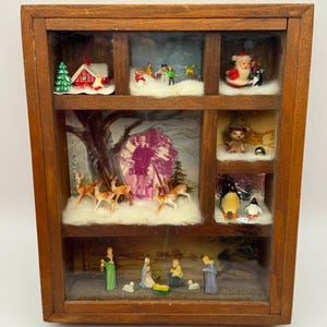 May include: A wooden shadow box with six compartments, each containing a miniature Christmas scene. Scenes include a snowy village, Santa Claus, reindeer, penguins, and a nativity scene. The box has a dark brown frame.