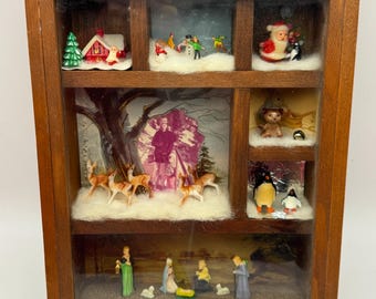 May include: A wooden shadow box with six compartments, each containing a miniature Christmas scene. Scenes include a snowy village, Santa Claus, reindeer, penguins, and a nativity scene. The box has a dark brown frame.