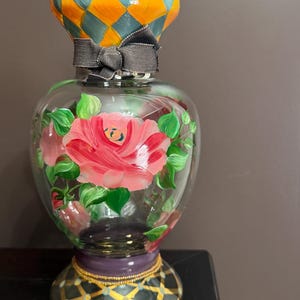 May include: A clear glass vase featuring hand-painted pink roses with green leaves on the body. The neck has an orange and teal diamond pattern, while the base displays a black and gold diamond design with a purple band. A grey ribbon is tied around the neck.