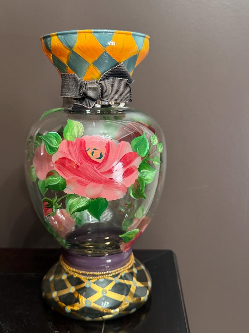 May include: A clear glass vase featuring hand-painted pink roses with green leaves on the body. The neck has an orange and teal diamond pattern, while the base displays a black and gold diamond design with a purple band. A grey ribbon is tied around the neck.