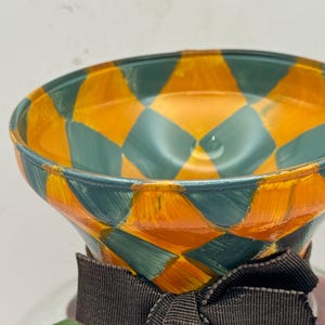 May include: A glass vessel featuring a flared rim painted with a vibrant orange and teal diamond pattern. A dark brown fabric ribbon is tied in a bow around the neck of the vessel, which sits above a base decorated with green leaf motifs.