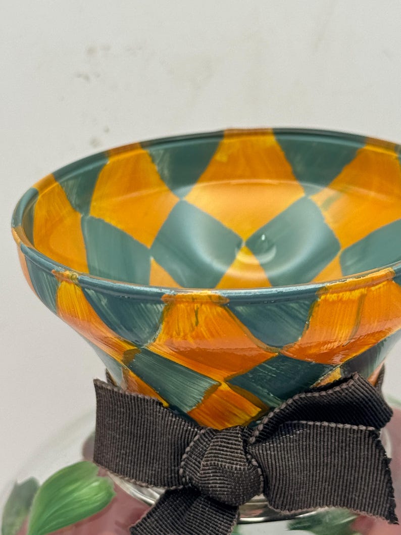May include: A glass vessel featuring a flared rim painted with a vibrant orange and teal diamond pattern. A dark brown fabric ribbon is tied in a bow around the neck of the vessel, which sits above a base decorated with green leaf motifs.