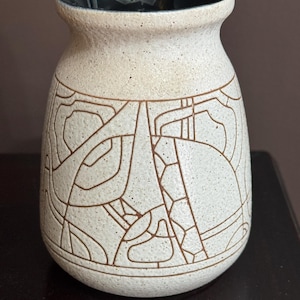 May include: A beige ceramic vase with a dark interior and a textured exterior. The vase features a brown geometric design wrapping around the body. The vase has a rounded base and a slightly flared rim. The vase is likely used for decorative purposes.
