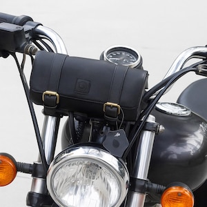 Pode incluir: Bolsa de guiador de motocicleta em couro preto com fivelas de latão. A bolsa cilíndrica é montada no guiador de uma motocicleta preta. A imagem mostra a frente da motocicleta, incluindo o farol e os piscas.