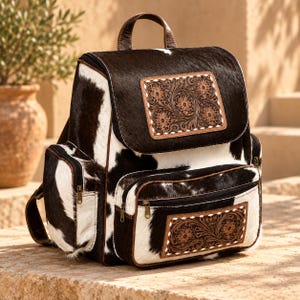 May include: A backpack crafted from black and white cowhide featuring a top handle and adjustable straps. The bag includes a front pocket and a main flap, both adorned with rectangular, brown leather panels featuring intricate floral tooling.
