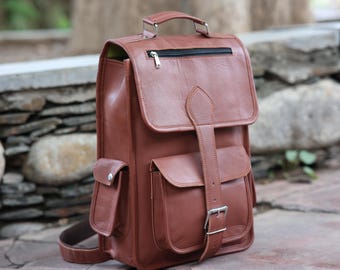 May include: A brown leather backpack with multiple pockets and a top handle. The bag features a flap closure with a buckle and a zippered top compartment. The backpack has adjustable shoulder straps and a small side pocket. The bag is made of a smooth, durable leather.