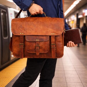 Puede incluir: Un maletín de cuero marrón con cierre de solapa, dos bolsillos delanteros y correas ajustables. La bolsa tiene herrajes de latón y un asa superior. Un cuaderno marrón se sostiene en la otra mano.