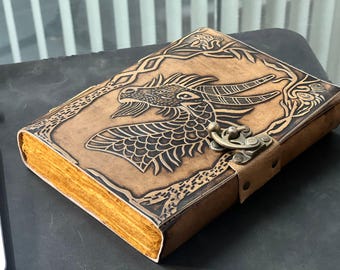 May include: A brown leather journal with a dragon design embossed on the cover. The journal has a metal clasp and aged paper pages. The dragon is intricately detailed, and the leather has a distressed appearance.
